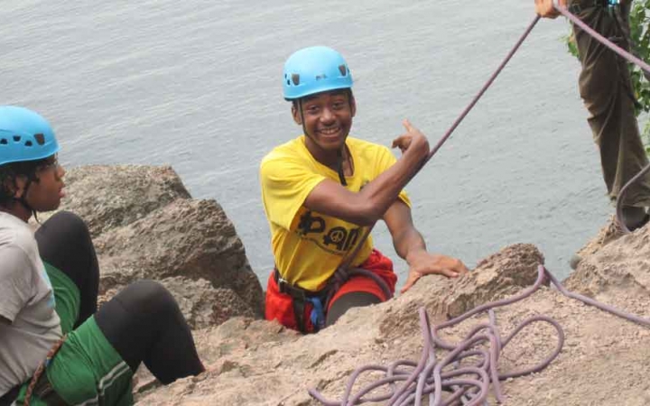 A person wearing safety gear is secured by ropes on the edge of a cliff high above a body of water and smiles excitedly as they point over their shoulder. 